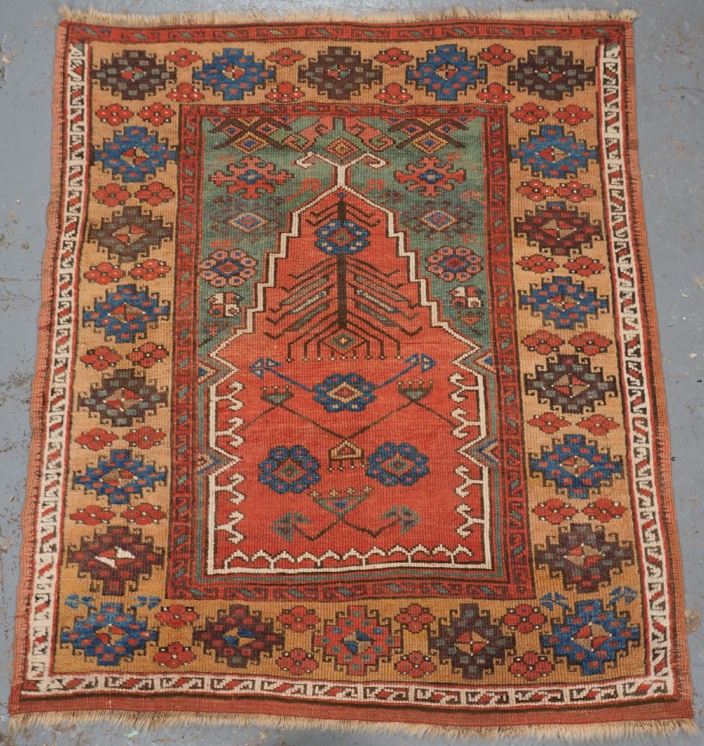 An antique hand-knotted Turkish Konya prayer rug featuring a stepped central red niche surrounded by green, blue, and ochre geometric motifs.