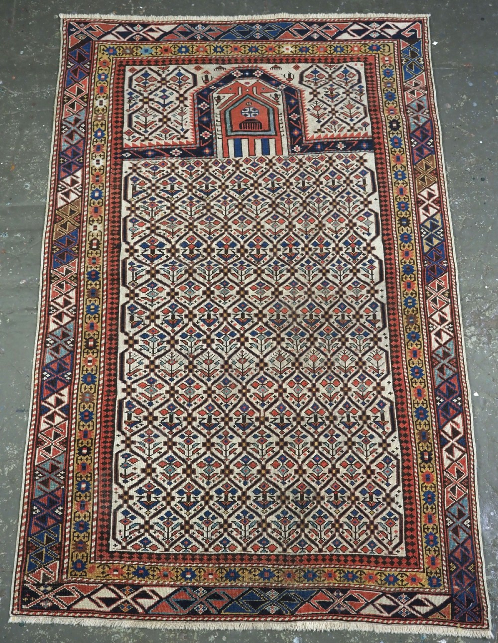 An antique Caucasian Shirvan wool prayer rug featuring an ivory field covered in a geometric floral lattice motif, enclosed by a multi-bordered frame and a stepped prayer arch at the top.