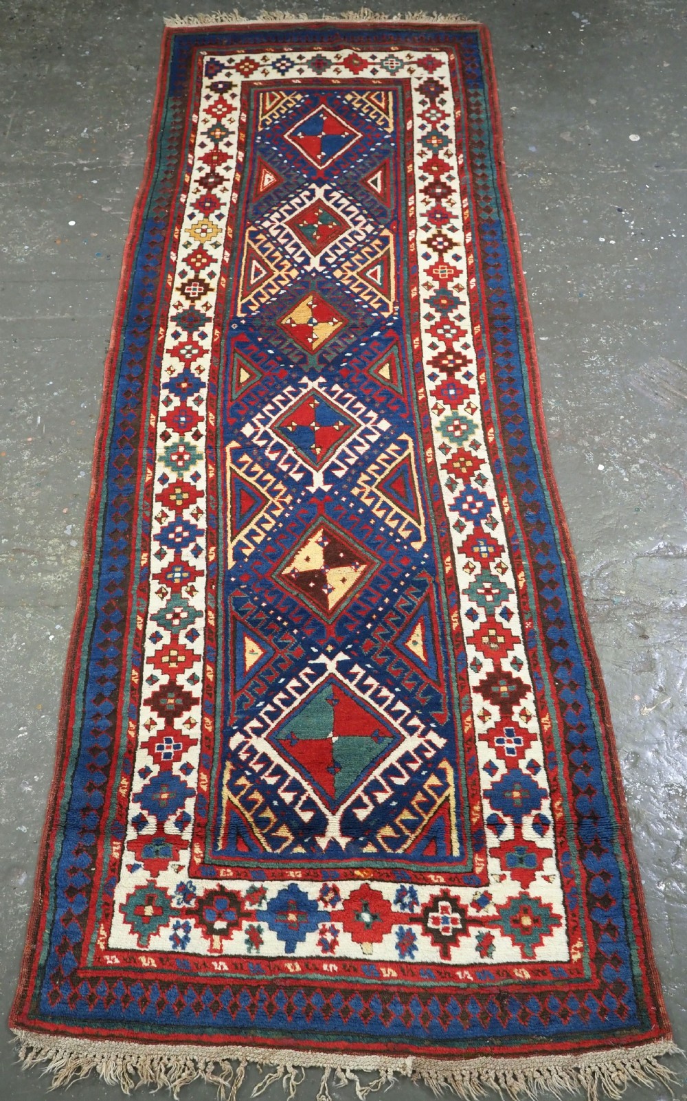 An antique Caucasian Moghan Kazak wool runner rug with repeating diamond hooked medallions on a deep blue field and geometric borders.