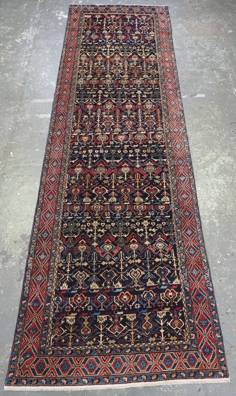 An antique handwoven wool Caucasian Kuba runner featuring dense geometric motifs in red, blue, and cream shades across a long dark navy field.