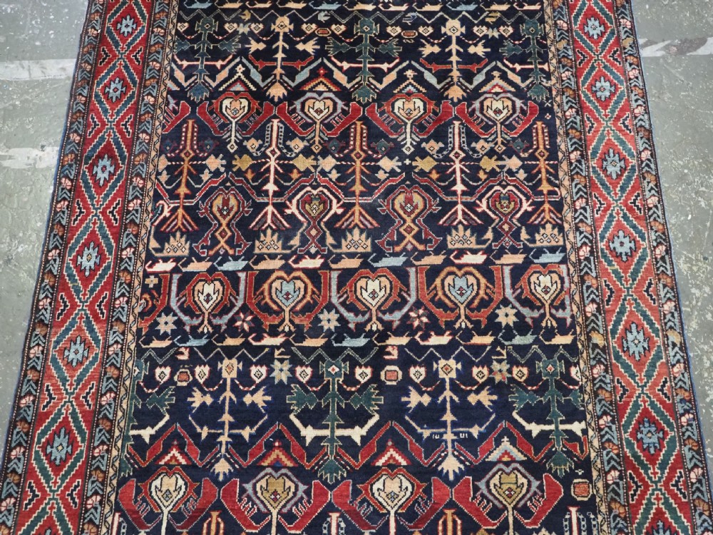 An antique handwoven wool Caucasian Kuba runner featuring dense geometric motifs in red, blue, and cream shades across a long dark navy field. View IV