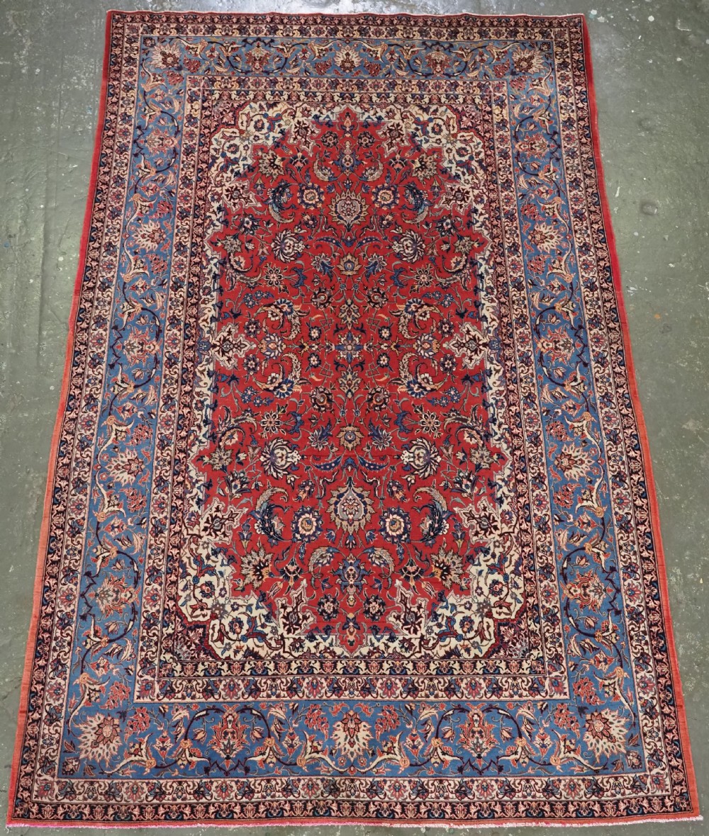 An antique Persian Isfahan carpet featuring a detailed red central field with floral motifs, framed by light spandrels and a wide blue floral border.
