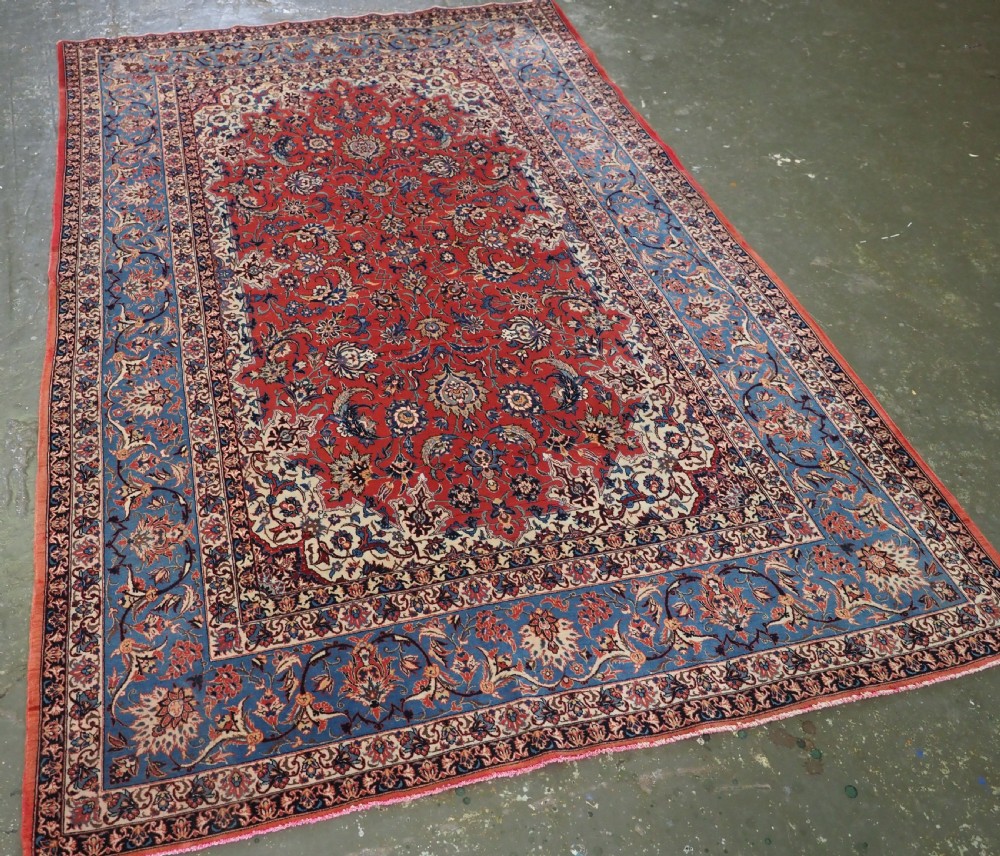 An antique Persian Isfahan carpet featuring a detailed red central field with floral motifs, framed by light spandrels and a wide blue floral border. View II