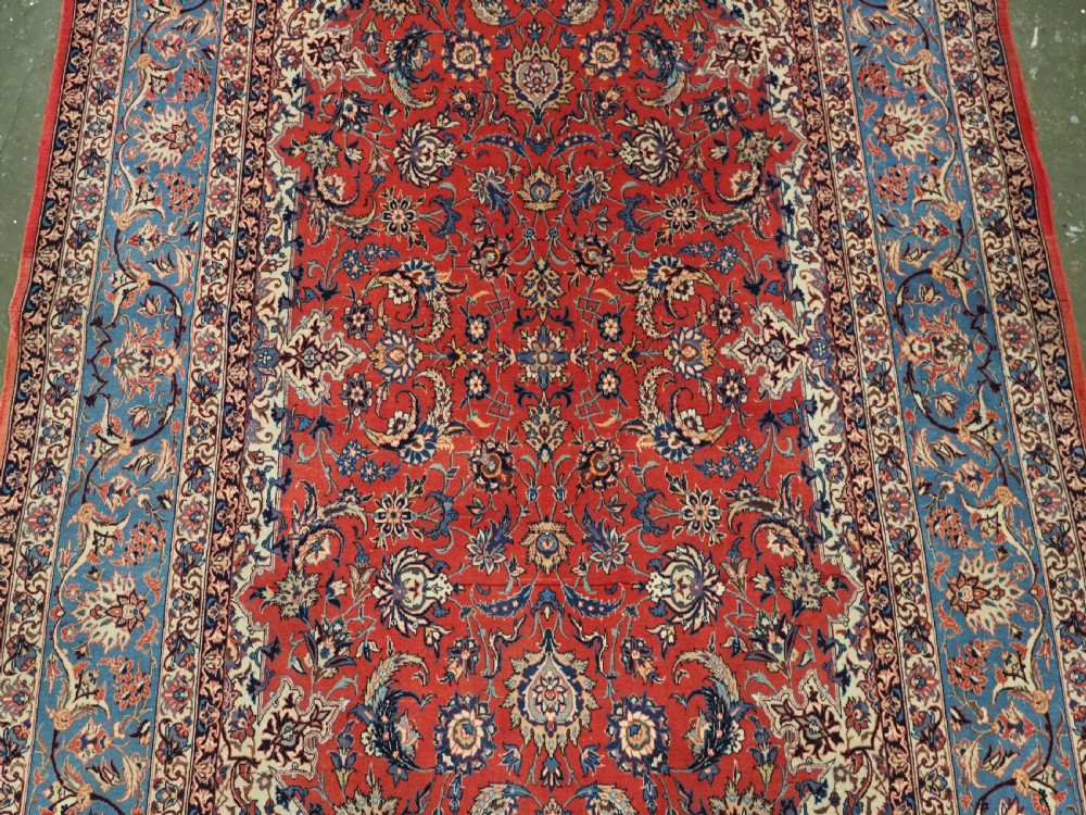 An antique Persian Isfahan carpet featuring a detailed red central field with floral motifs, framed by light spandrels and a wide blue floral border. View III