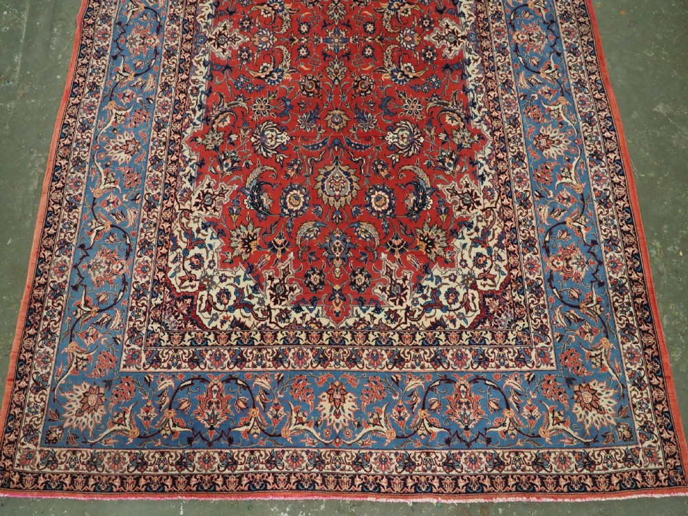 An antique Persian Isfahan carpet featuring a detailed red central field with floral motifs, framed by light spandrels and a wide blue floral border. View IV