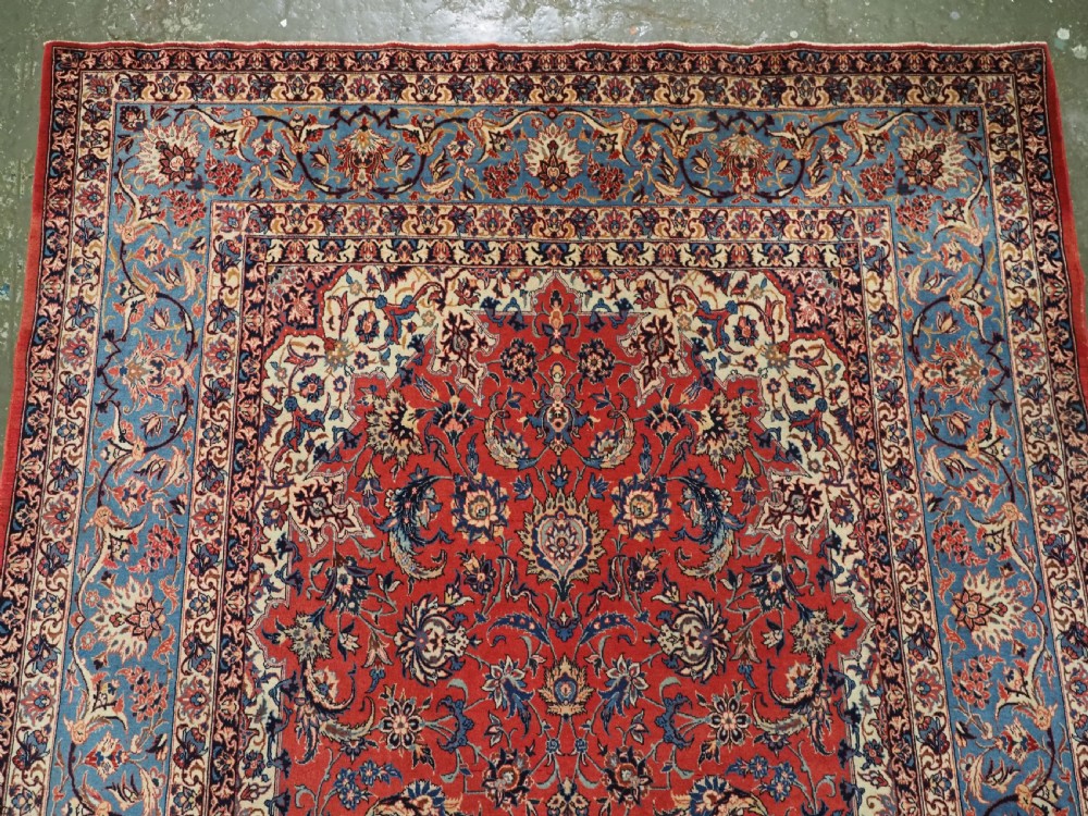 An antique Persian Isfahan carpet featuring a detailed red central field with floral motifs, framed by light spandrels and a wide blue floral border. View V