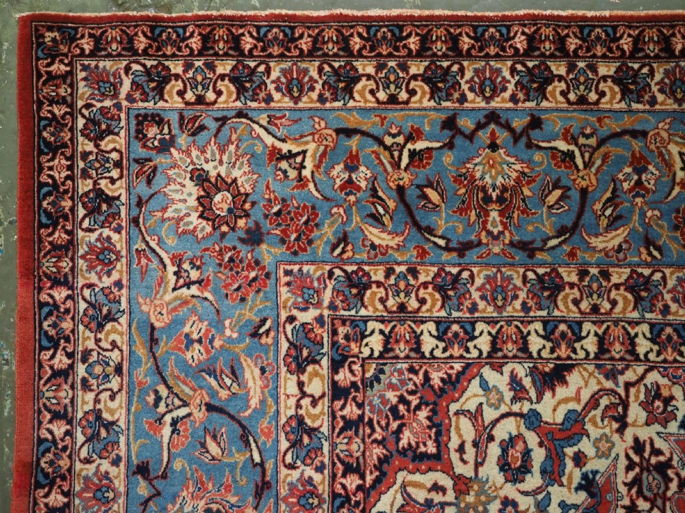 An antique Persian Isfahan carpet featuring a detailed red central field with floral motifs, framed by light spandrels and a wide blue floral border. View VI