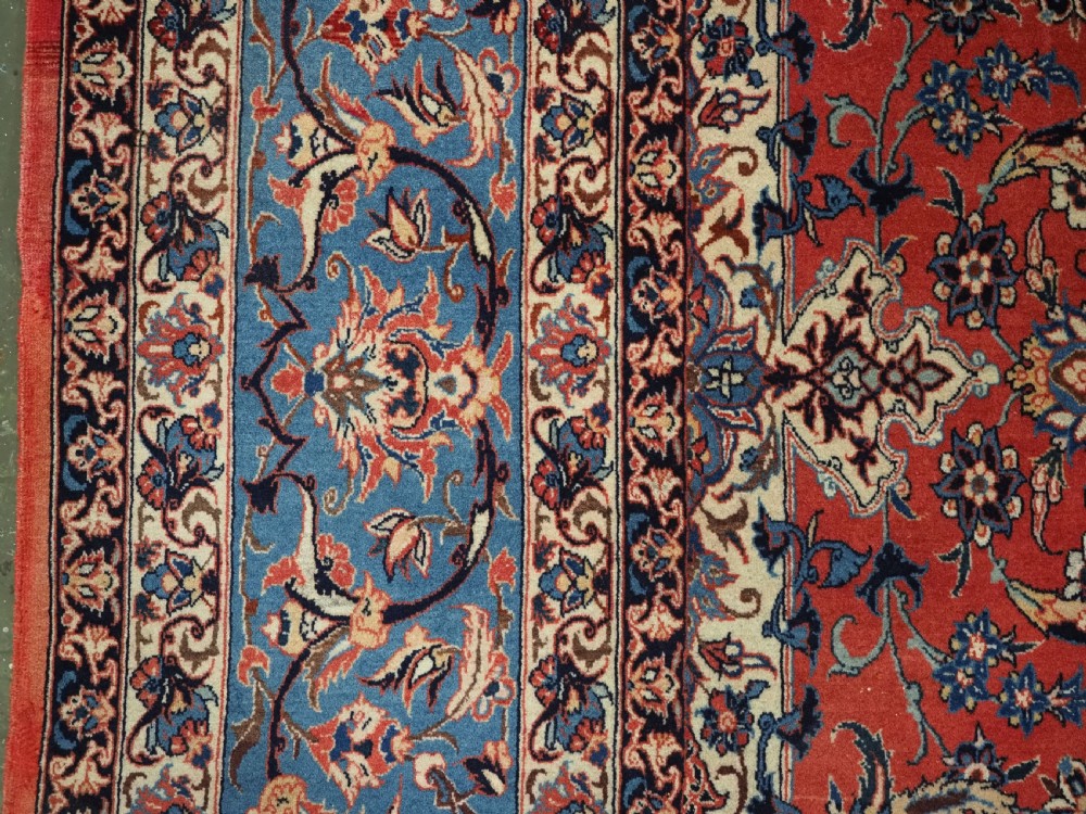 An antique Persian Isfahan carpet featuring a detailed red central field with floral motifs, framed by light spandrels and a wide blue floral border. View VII