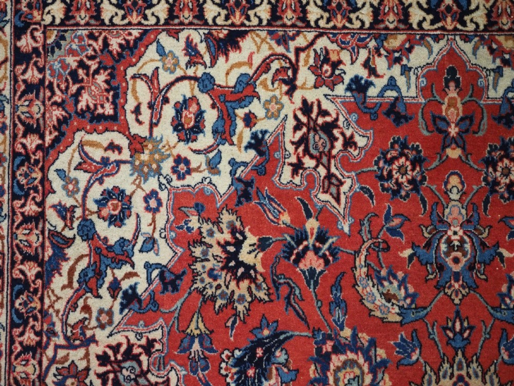 An antique Persian Isfahan carpet featuring a detailed red central field with floral motifs, framed by light spandrels and a wide blue floral border. View VIII