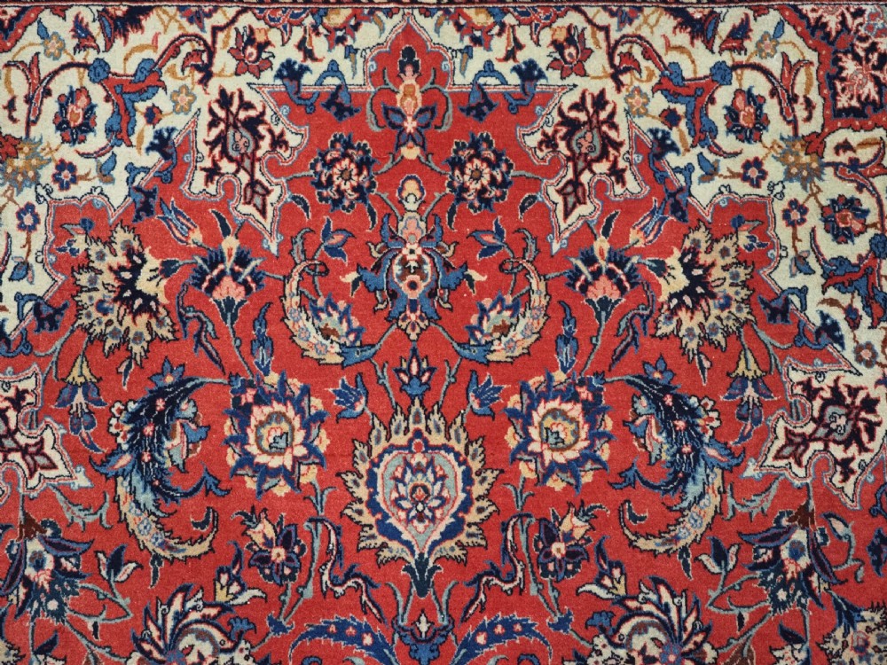 An antique Persian Isfahan carpet featuring a detailed red central field with floral motifs, framed by light spandrels and a wide blue floral border. View IX