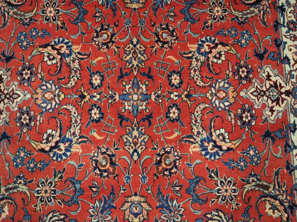 An antique Persian Isfahan carpet featuring a detailed red central field with floral motifs, framed by light spandrels and a wide blue floral border. View X
