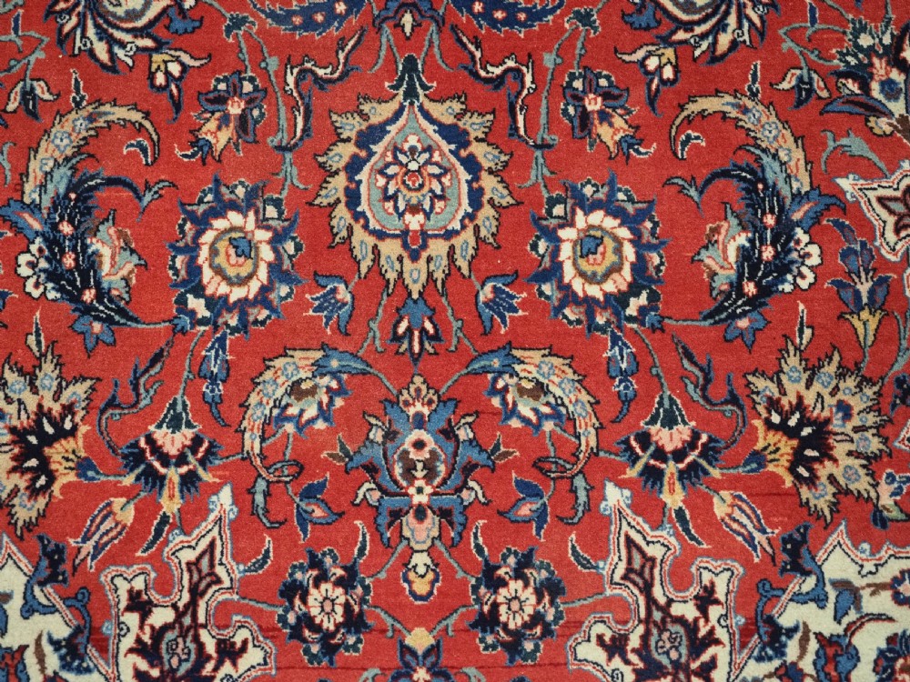 An antique Persian Isfahan carpet featuring a detailed red central field with floral motifs, framed by light spandrels and a wide blue floral border. View XI