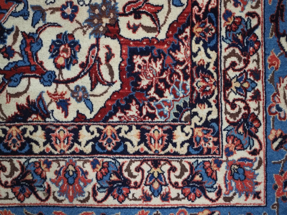 An antique Persian Isfahan carpet featuring a detailed red central field with floral motifs, framed by light spandrels and a wide blue floral border. View XIII