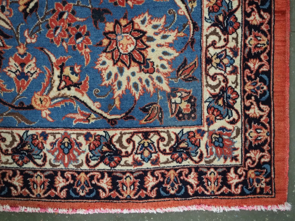 An antique Persian Isfahan carpet featuring a detailed red central field with floral motifs, framed by light spandrels and a wide blue floral border. View XIV