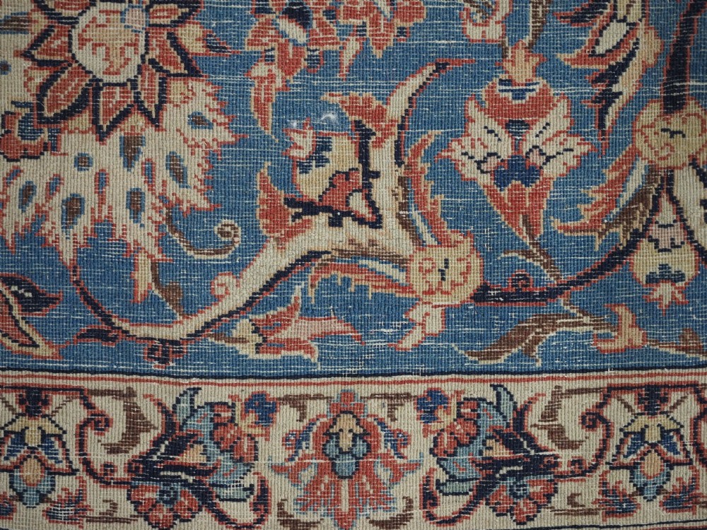 An antique Persian Isfahan carpet featuring a detailed red central field with floral motifs, framed by light spandrels and a wide blue floral border. View XV