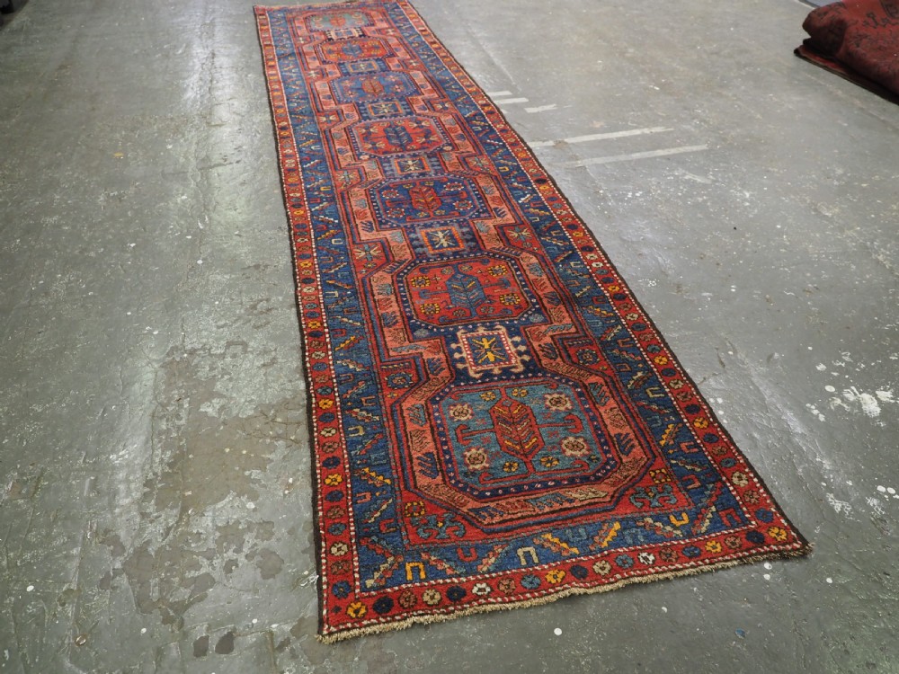 A long hand-knotted wool runner rug featuring geometric garden medallions in indigo blue, terracotta red, and gold within a patterned border. View II