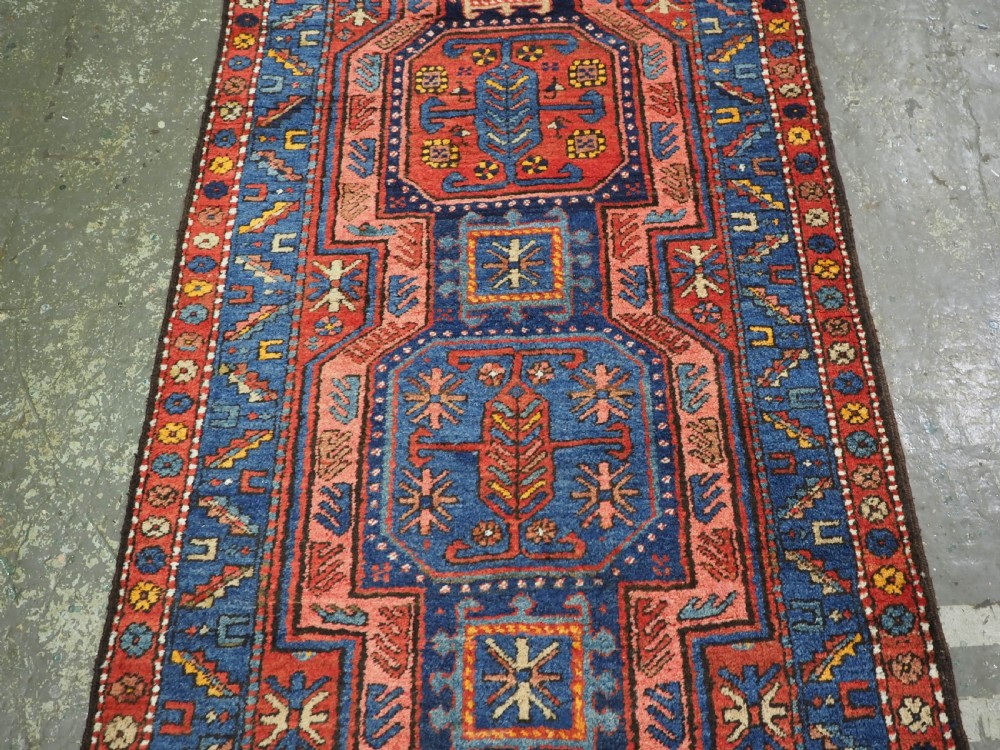 A long hand-knotted wool runner rug featuring geometric garden medallions in indigo blue, terracotta red, and gold within a patterned border. View V
