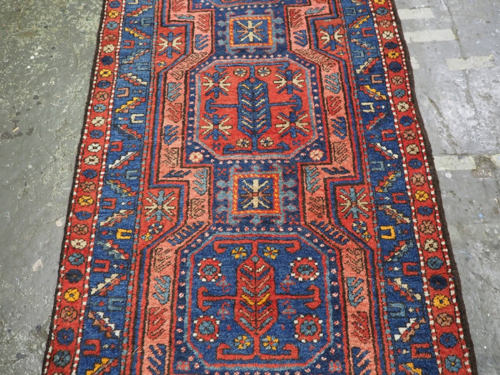 A long hand-knotted wool runner rug featuring geometric garden medallions in indigo blue, terracotta red, and gold within a patterned border. View VI