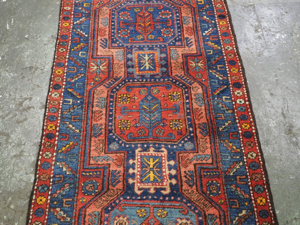 A long hand-knotted wool runner rug featuring geometric garden medallions in indigo blue, terracotta red, and gold within a patterned border. View VII