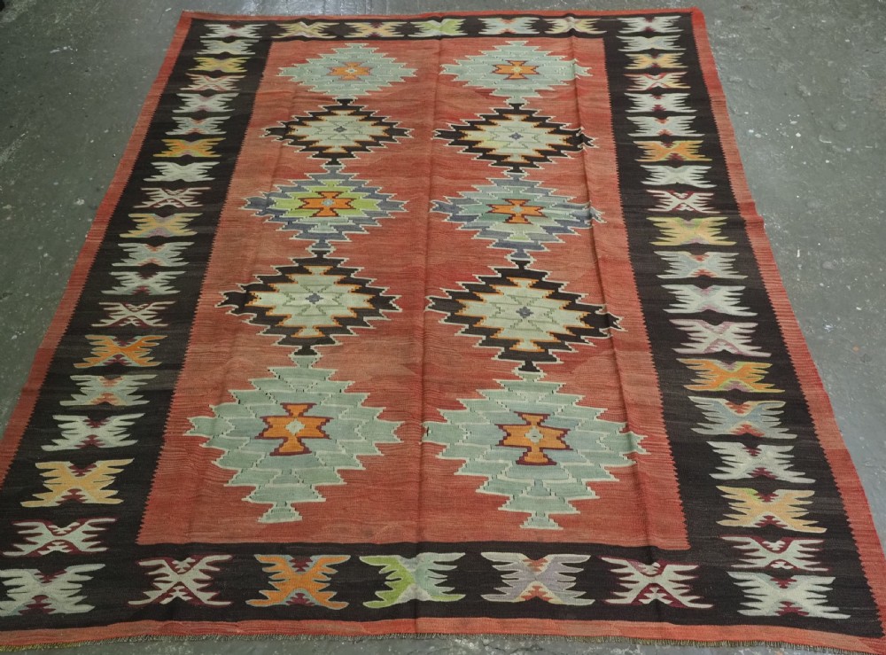 A flat-woven wool kilim rug featuring a terracotta red field with repeating sage green and cream stepped medallions framed by a dark brown geometric border.