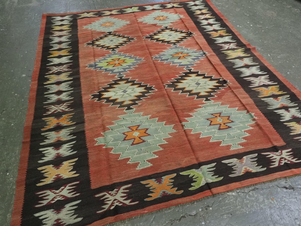 A flat-woven wool kilim rug featuring a terracotta red field with repeating sage green and cream stepped medallions framed by a dark brown geometric border. View II