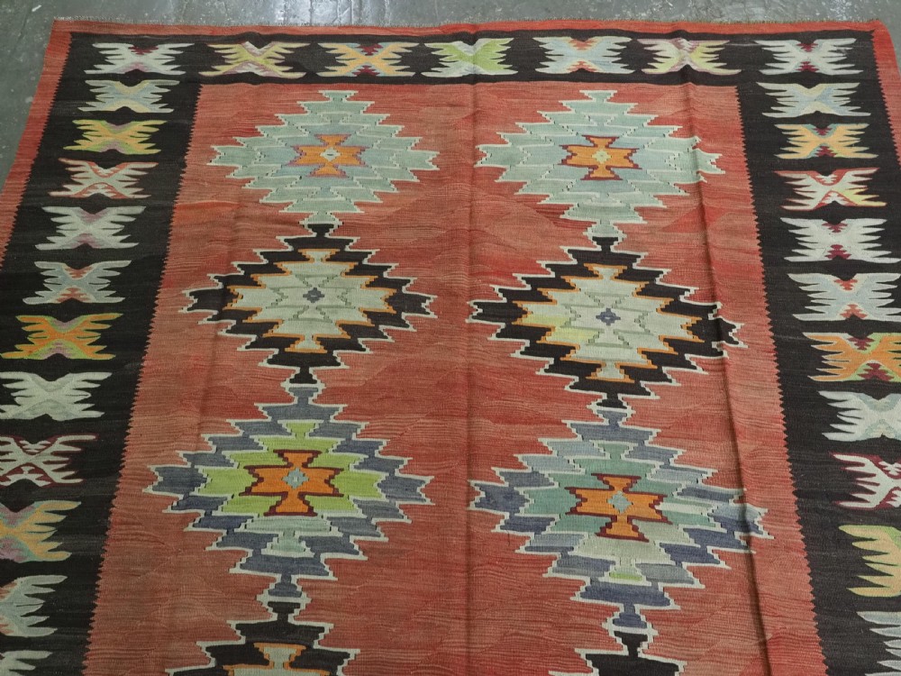 A flat-woven wool kilim rug featuring a terracotta red field with repeating sage green and cream stepped medallions framed by a dark brown geometric border. View III
