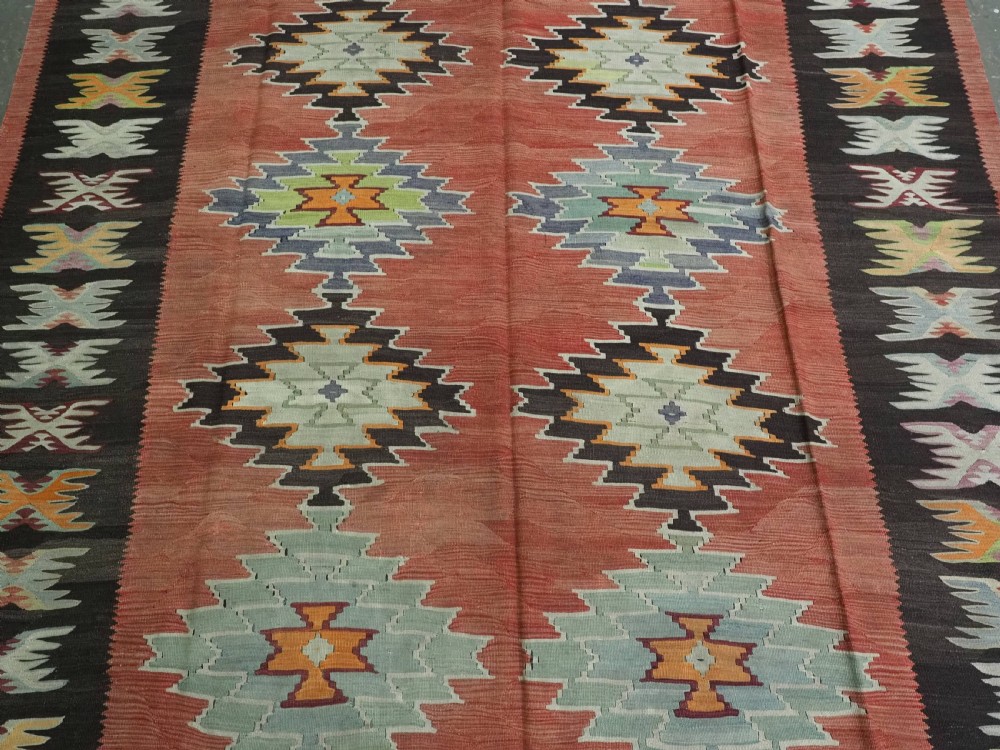 A flat-woven wool kilim rug featuring a terracotta red field with repeating sage green and cream stepped medallions framed by a dark brown geometric border. View IV