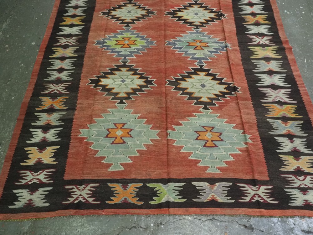 A flat-woven wool kilim rug featuring a terracotta red field with repeating sage green and cream stepped medallions framed by a dark brown geometric border. View V