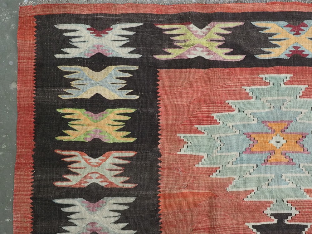 A flat-woven wool kilim rug featuring a terracotta red field with repeating sage green and cream stepped medallions framed by a dark brown geometric border. View VI