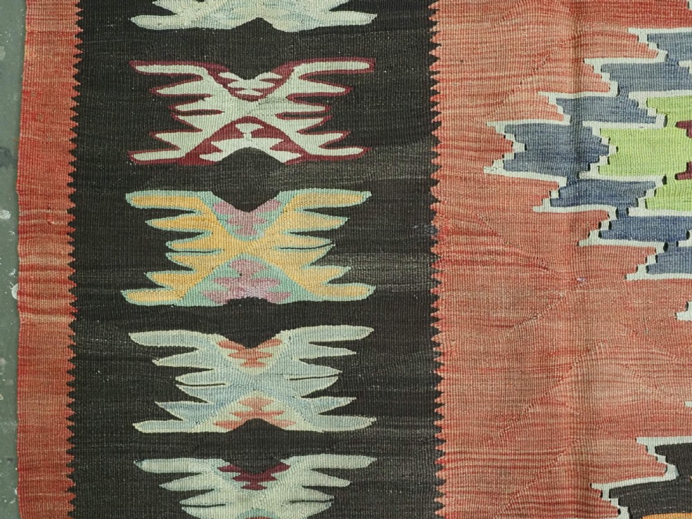 A flat-woven wool kilim rug featuring a terracotta red field with repeating sage green and cream stepped medallions framed by a dark brown geometric border. View VII