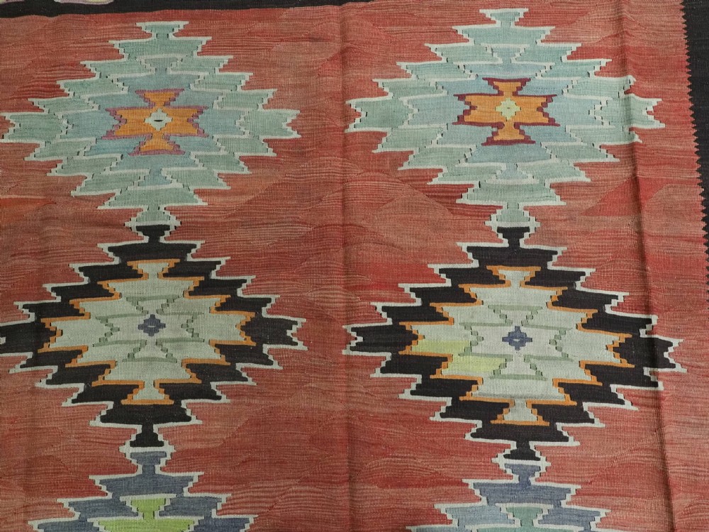 A flat-woven wool kilim rug featuring a terracotta red field with repeating sage green and cream stepped medallions framed by a dark brown geometric border. View VIII