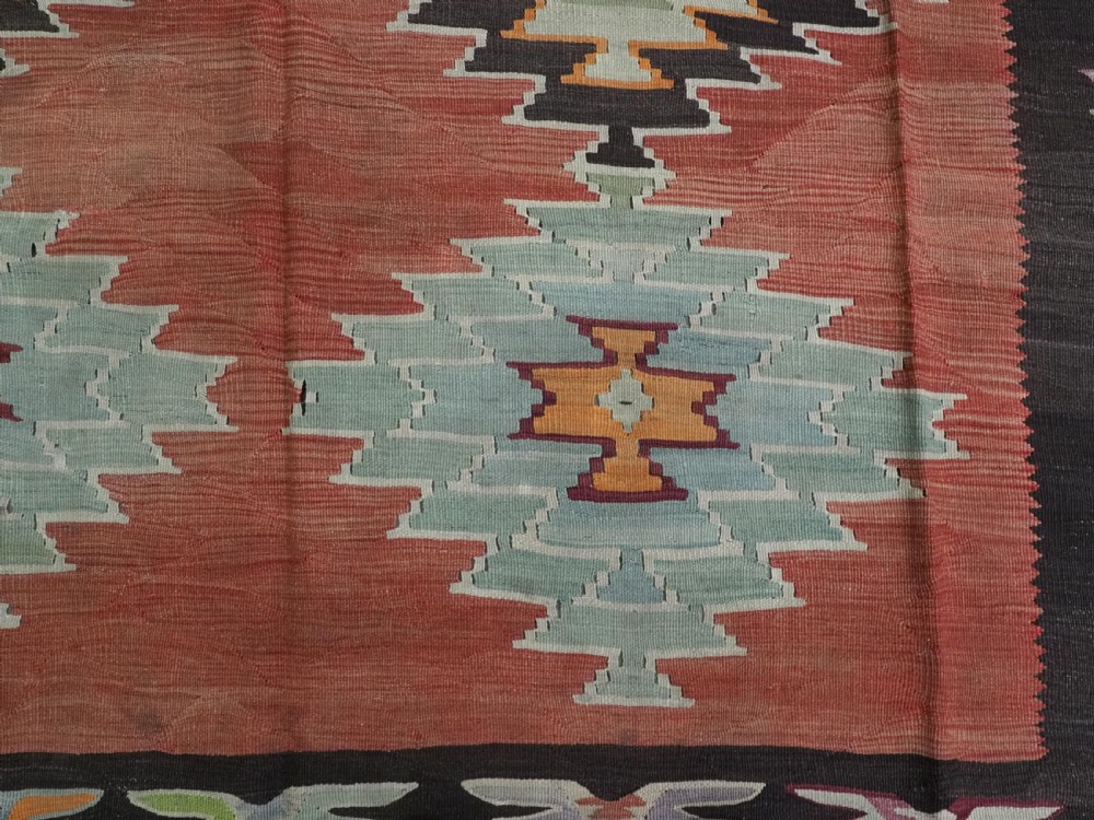 A flat-woven wool kilim rug featuring a terracotta red field with repeating sage green and cream stepped medallions framed by a dark brown geometric border. View IX