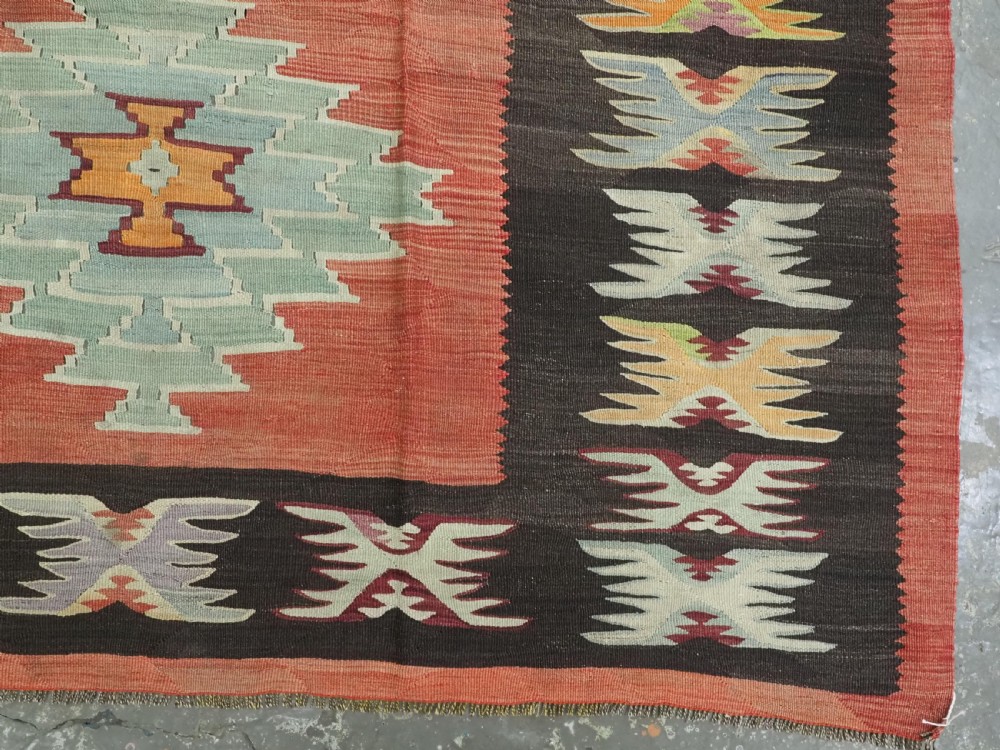 A flat-woven wool kilim rug featuring a terracotta red field with repeating sage green and cream stepped medallions framed by a dark brown geometric border. View X