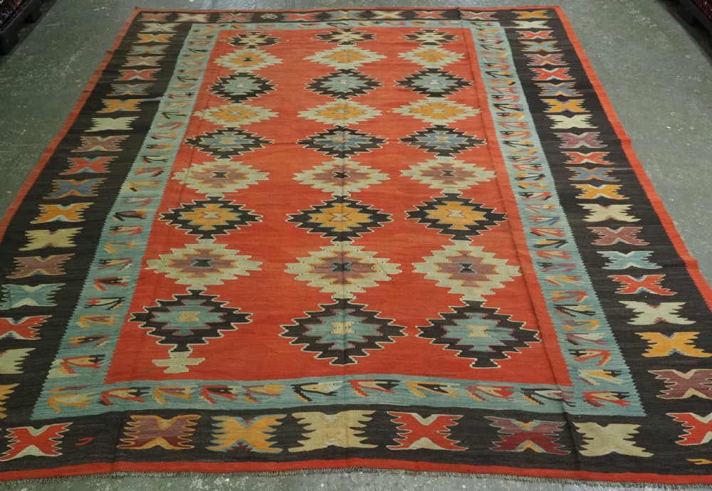A large antique Pirot Sarkoy flat-weave wool kilim carpet featuring repeating diamond medallions on a terracotta field with dark geometric borders.