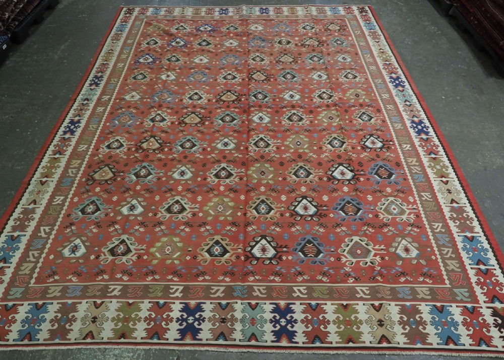 An antique handwoven wool kilim rug featuring a terracotta central field with geometric repeat motifs and patterned borders.