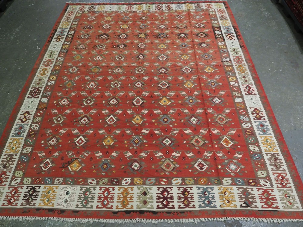A large antique Pirot Sarkoy flatweave wool kilim rug featuring repeating geometric diamond motifs on a terracotta red field with decorative patterned borders.