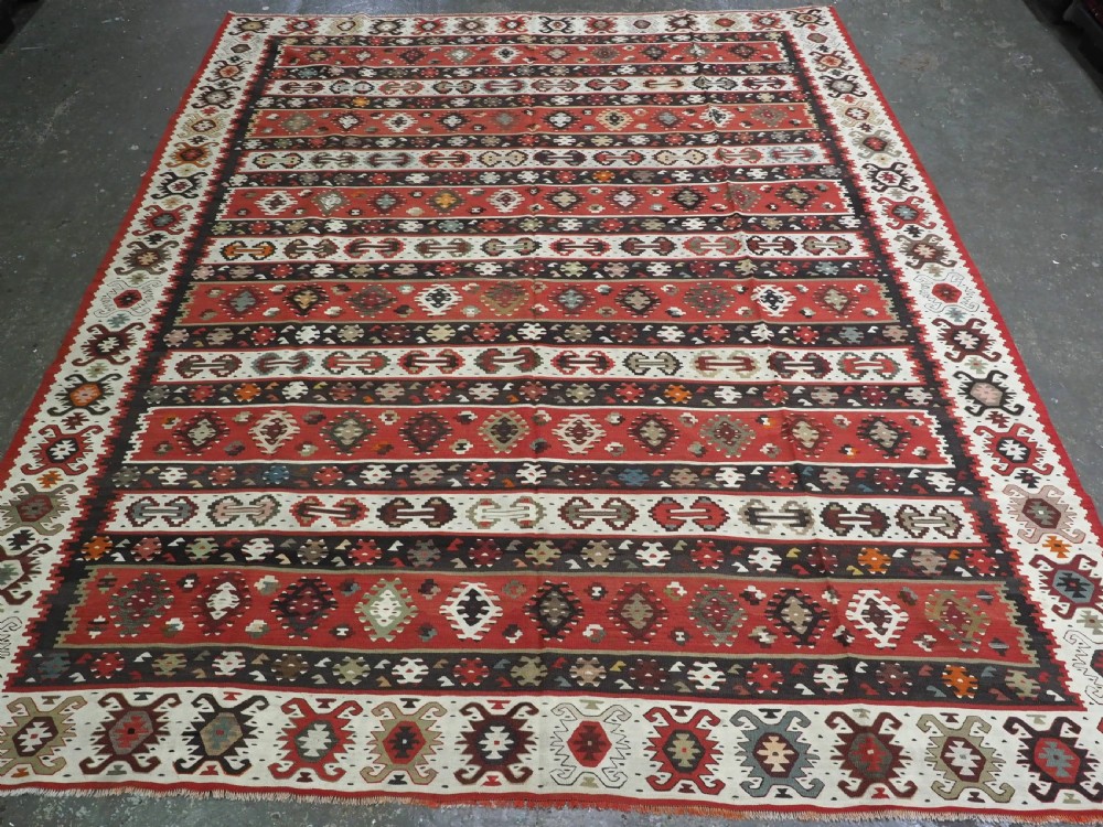 A large handwoven antique kilim rug featuring horizontal bands of intricate geometric motifs in terracotta red, cream, charcoal, and brown hues.