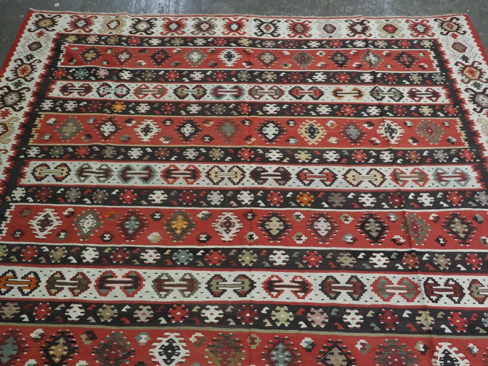 A large handwoven antique kilim rug featuring horizontal bands of intricate geometric motifs in terracotta red, cream, charcoal, and brown hues. View III