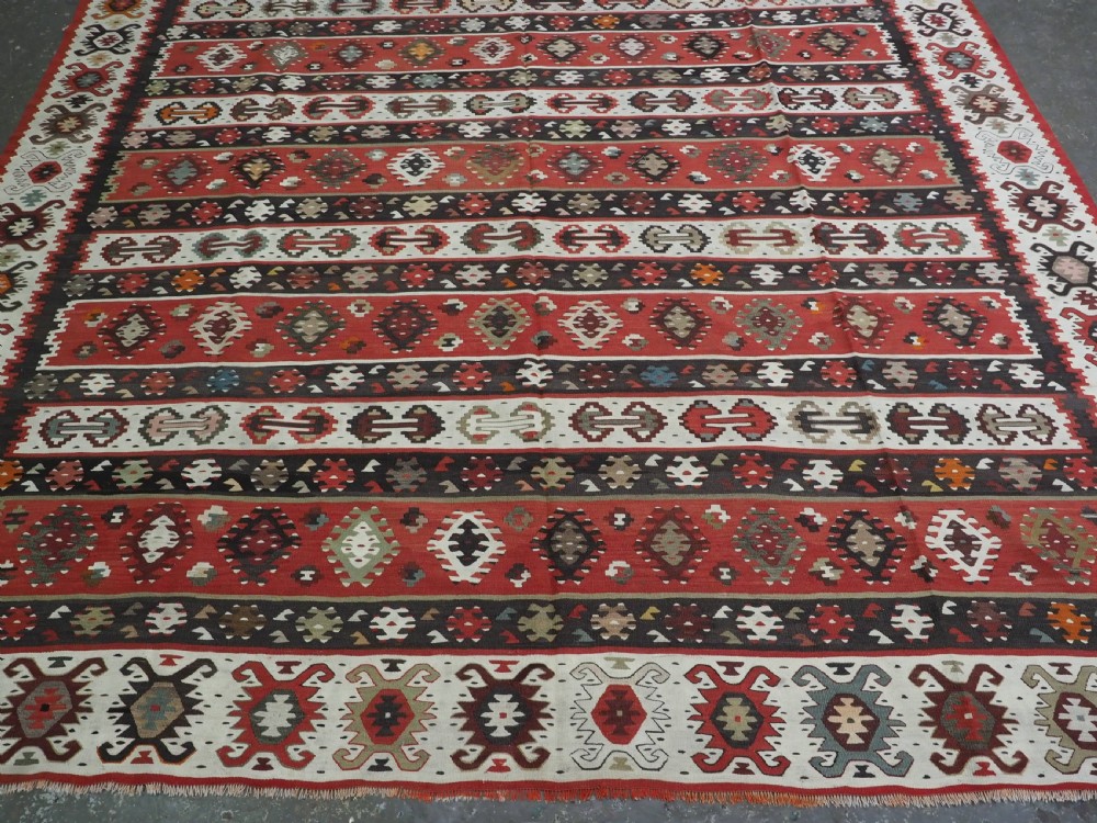 A large handwoven antique kilim rug featuring horizontal bands of intricate geometric motifs in terracotta red, cream, charcoal, and brown hues. View IV