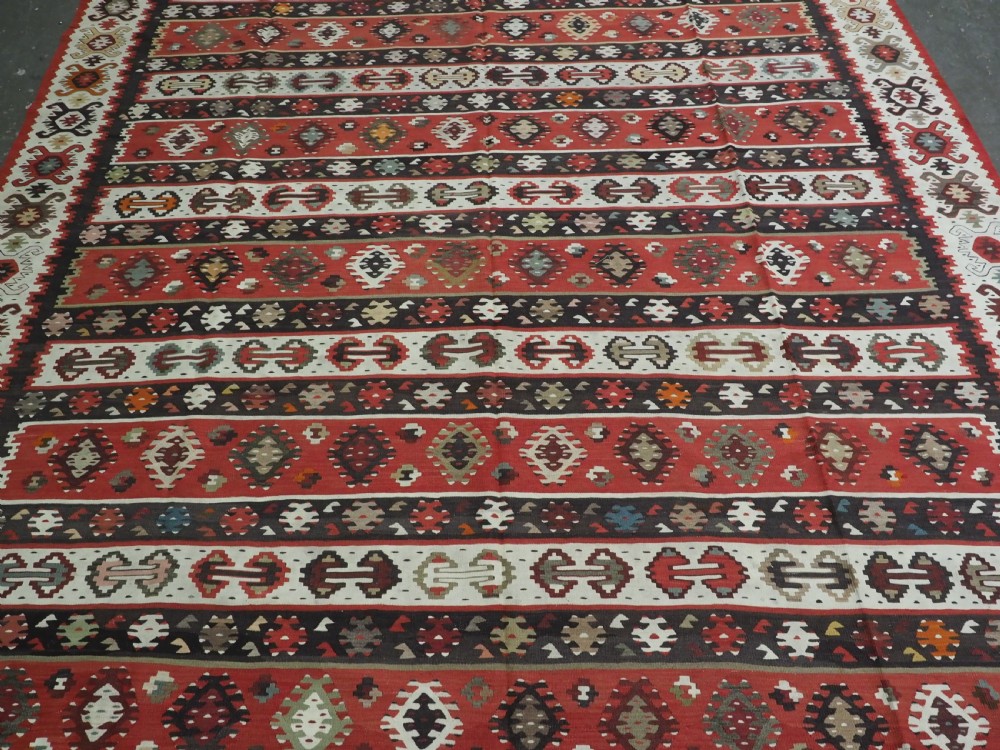 A large handwoven antique kilim rug featuring horizontal bands of intricate geometric motifs in terracotta red, cream, charcoal, and brown hues. View V