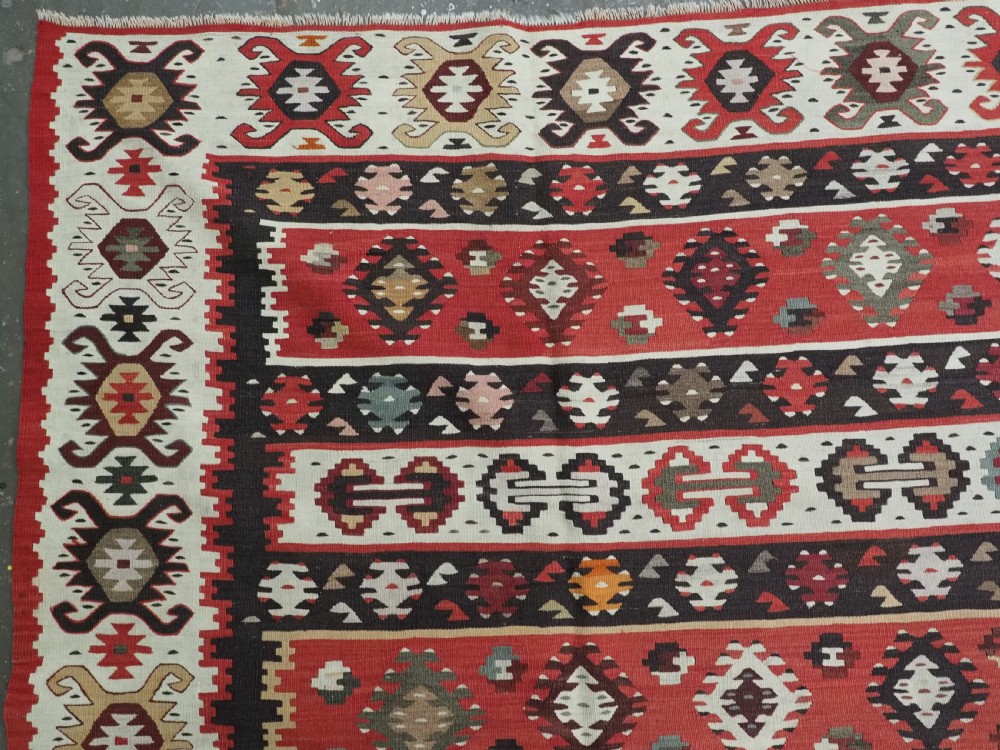 A large handwoven antique kilim rug featuring horizontal bands of intricate geometric motifs in terracotta red, cream, charcoal, and brown hues. View VI