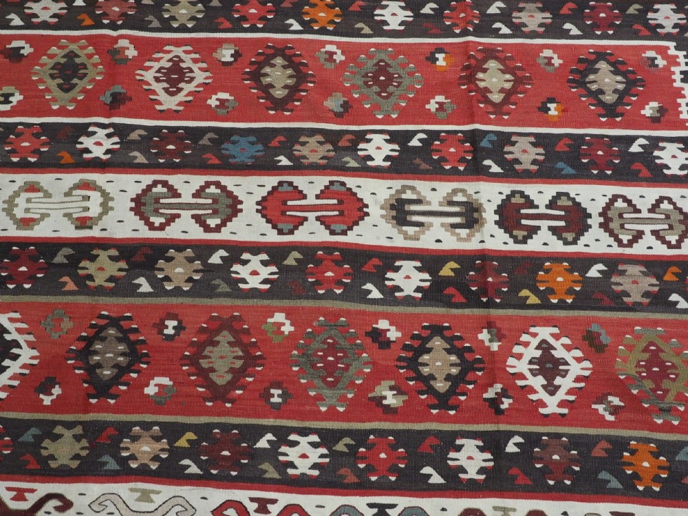 A large handwoven antique kilim rug featuring horizontal bands of intricate geometric motifs in terracotta red, cream, charcoal, and brown hues. View X