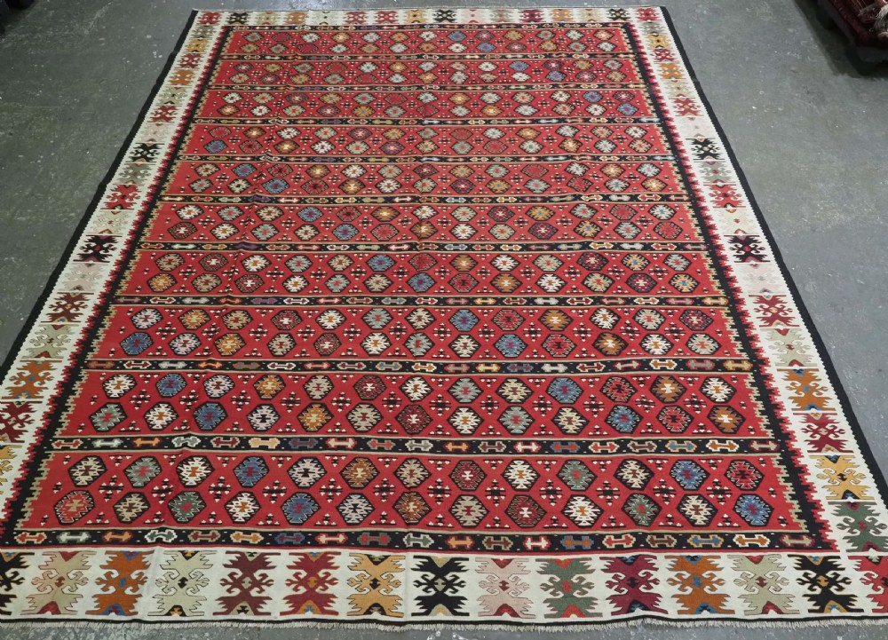 A large antique red flatweave wool kilim rug featuring horizontal bands filled with geometric diamond patterns and detailed multicoloured borders.