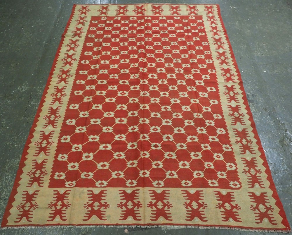 A rectangular flatweave wool kilim carpet featuring a red centre field with a cream lattice pattern, enclosed by a geometric cream and red border.