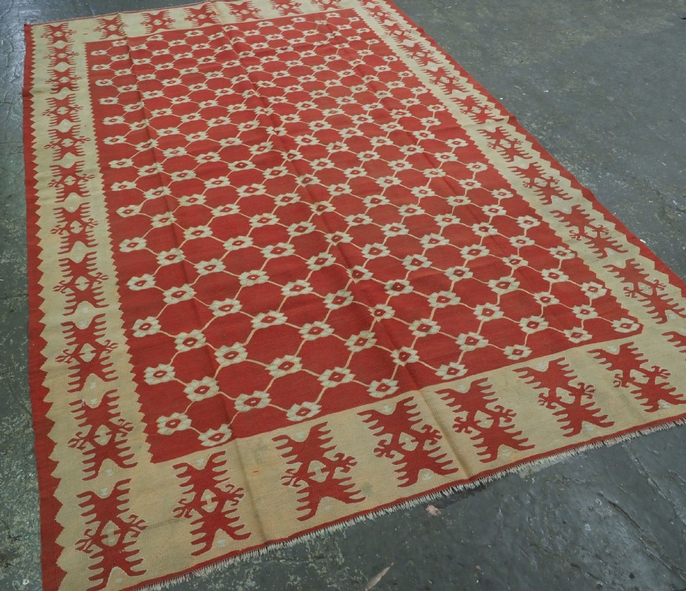 A rectangular flatweave wool kilim carpet featuring a red centre field with a cream lattice pattern, enclosed by a geometric cream and red border. View II