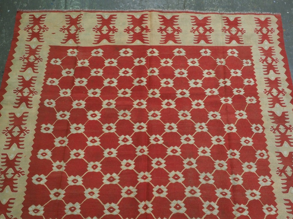 A rectangular flatweave wool kilim carpet featuring a red centre field with a cream lattice pattern, enclosed by a geometric cream and red border. View III