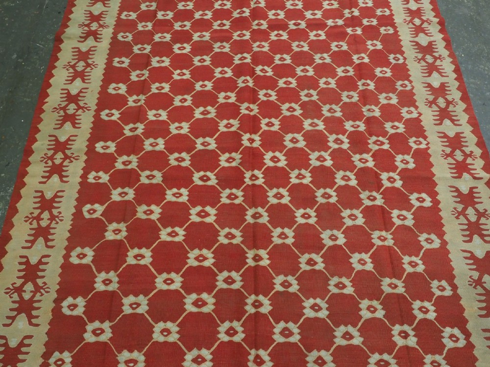 A rectangular flatweave wool kilim carpet featuring a red centre field with a cream lattice pattern, enclosed by a geometric cream and red border. View IV
