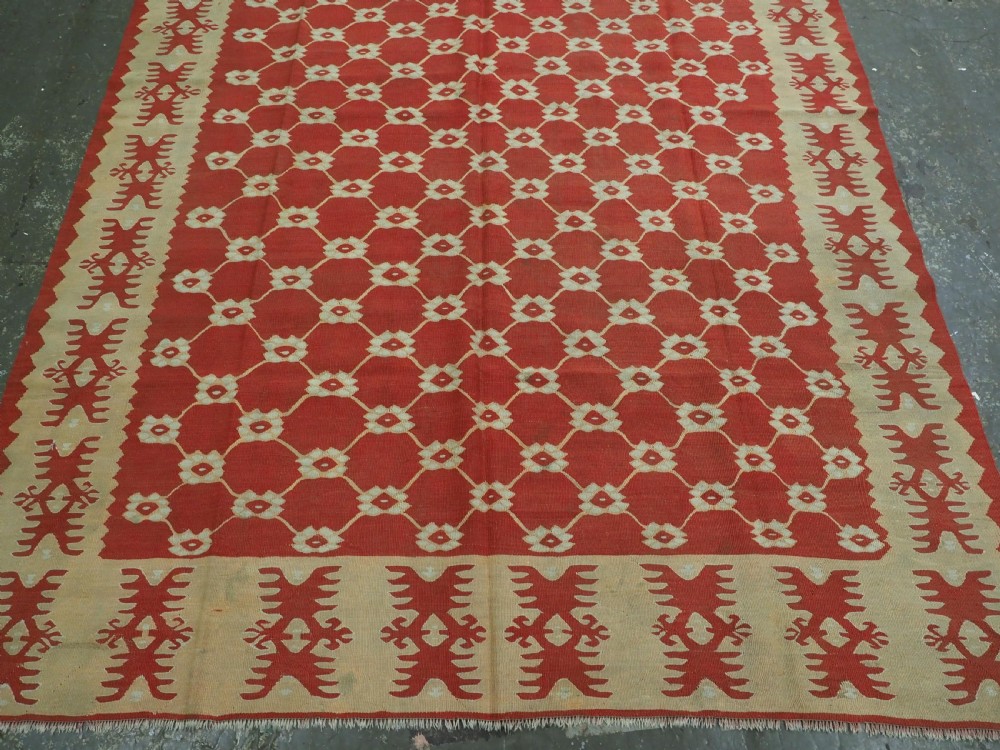 A rectangular flatweave wool kilim carpet featuring a red centre field with a cream lattice pattern, enclosed by a geometric cream and red border. View V