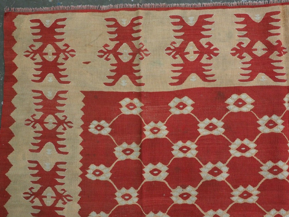 A rectangular flatweave wool kilim carpet featuring a red centre field with a cream lattice pattern, enclosed by a geometric cream and red border. View VI
