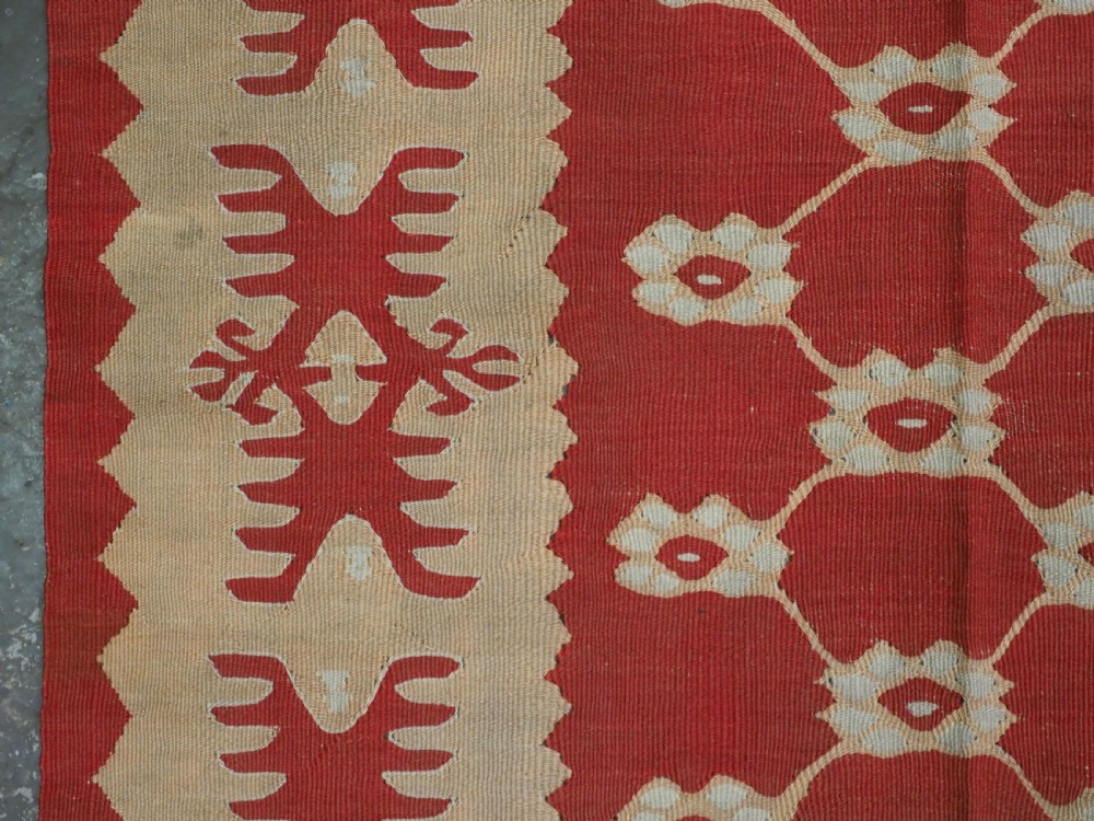 A rectangular flatweave wool kilim carpet featuring a red centre field with a cream lattice pattern, enclosed by a geometric cream and red border. View VII